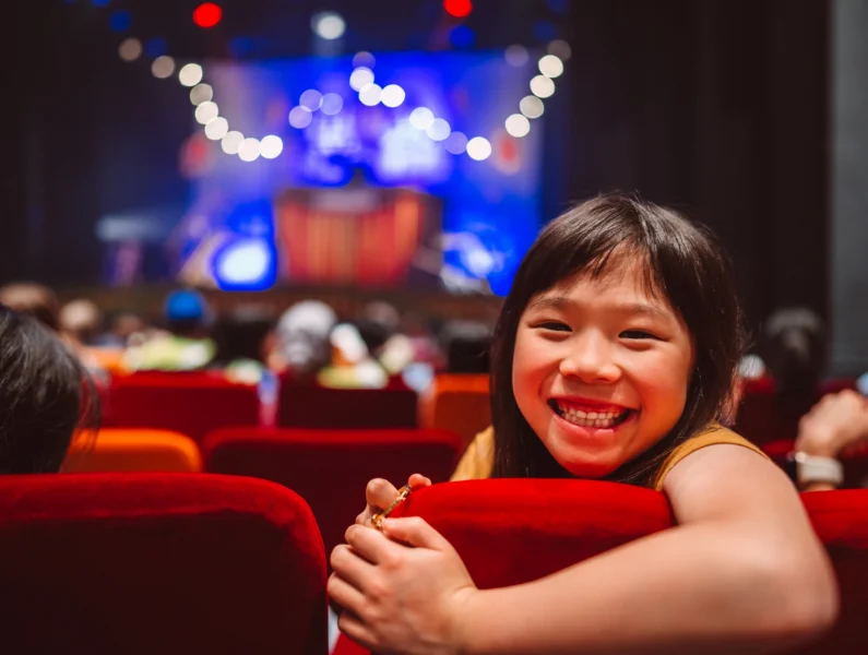 criança sentada numa sala de espetáculos, entusiasmada antes de um concerto ao espetáculo infantil.
