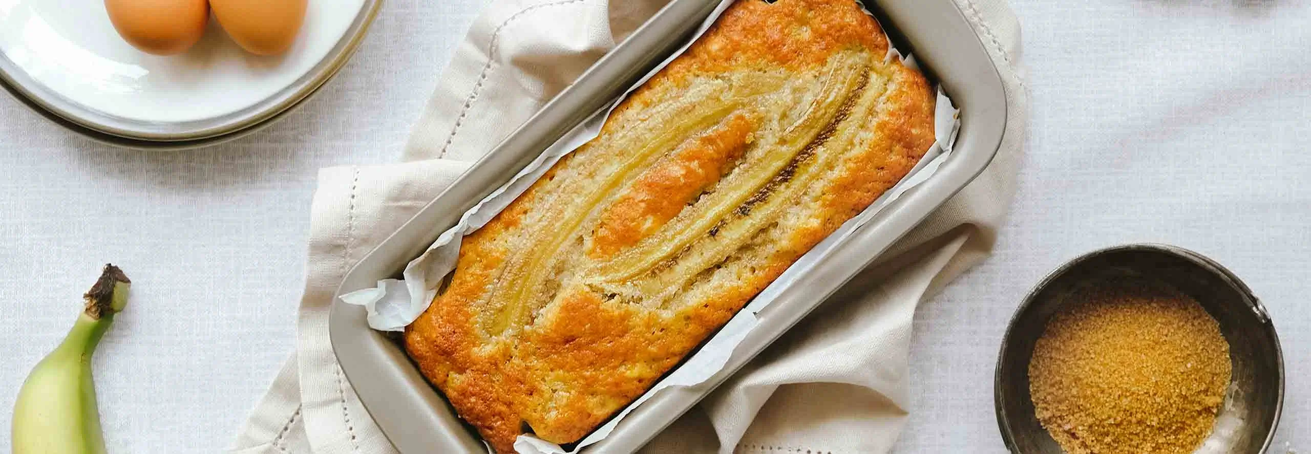 Pão de banana acabado de sair do forno sobre uma tábua de madeira, rodeado dos ingredientes frescos usados para a receita.