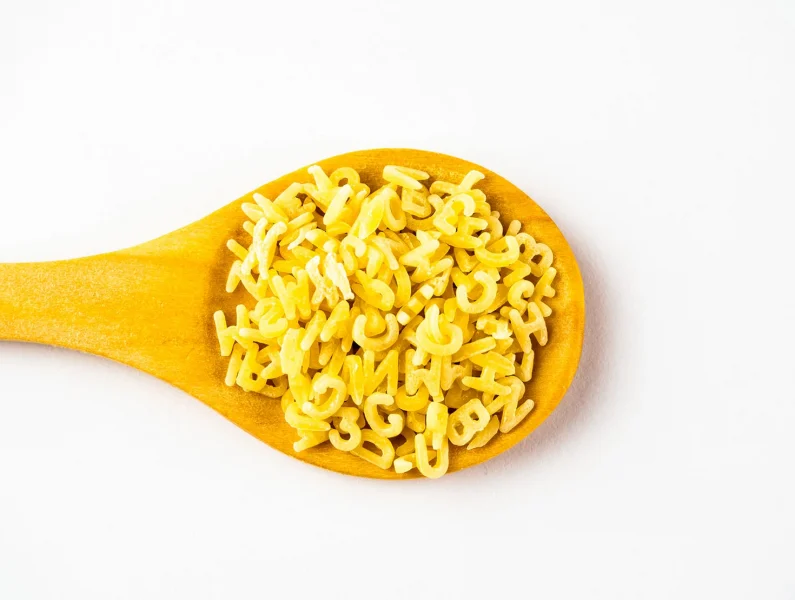 Vista superior de uma colher de pau com massa de sopa de letrinhas crua, antes da cozedura.
