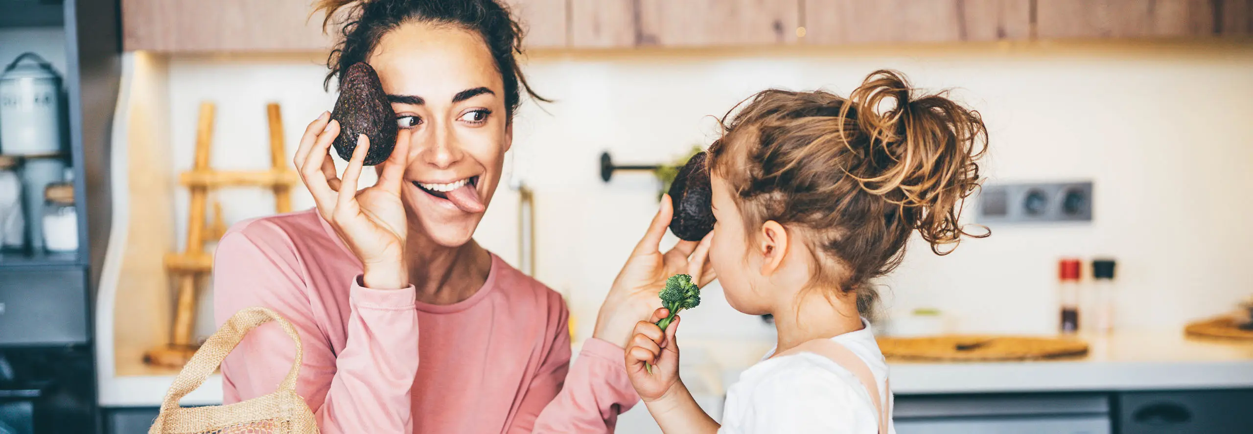 Mãe e filha divertidas numa cozinha, rodeadas de legumes frescos.